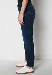 Jeans de mezclilla oscura con un diseño de pierna recta. Estilo de cinco bolsillos con cintura alta y detalles de costura sutiles, combinados con zapatillas blancas.
