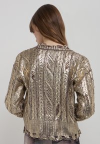 Pull en or métallique avec un motif en maille torsadée texturée, manches longues et ourlet court. Des sections transparentes ajoutent de la profondeur au design.