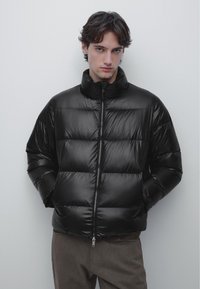 Młody mężczyzna z rękami w kieszeniach, ubrany w czarną pikowaną kurtkę i brązowe spodnie, stoi na tle jednolitego jasnoszarego tła.