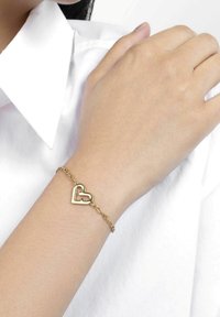 Braccialetto in oro con design a forma di cuore. Finitura liscia e lucida con un accento a forma di cuore aperto. Indossato su un polso di tonalità della pelle chiara.