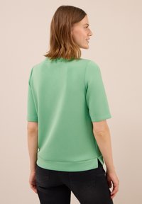 Femme à cheveux bruns longs jusqu'aux épaules, portant un haut vert menthe à manches courtes et un jean noir, de profil contre un fond beige.