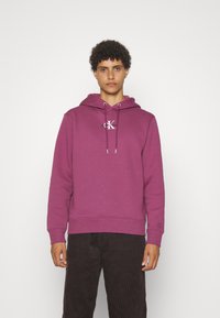 Hoodie di cotone viola con tasca frontale, cappuccio con cordino e logo sul petto, abbinato a pantaloni di velluto a coste scuri.
