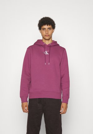 Hoodie di cotone viola con tasca frontale, cappuccio con cordino e logo sul petto, abbinato a pantaloni di velluto a coste scuri.