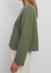 Pull vert tricoté avec un col rond, coupe courte et poignets côtelés. Texture douce et coupe ample, accompagné d'un pantalon de couleur claire.