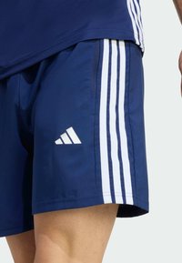 Tmavomodré športové šortky s bielymi tromi pruhmi a bielym logom Adidas na spodnej ľavej strane. Hladká textúra látky.