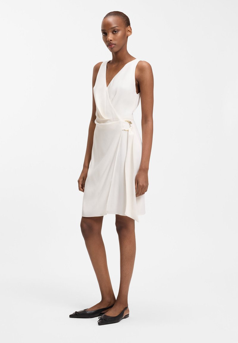 Femme portant une robe portefeuille blanche sans manches avec un détail de boucle dorée, associée à des ballerines noires à bouts pointus, debout devant un fond blanc uni.