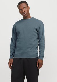 Hombre vestido con un sudadera de cuello redondo azul grisáceo de manga larga y pantalones negros, de pie contra un fondo blanco liso.