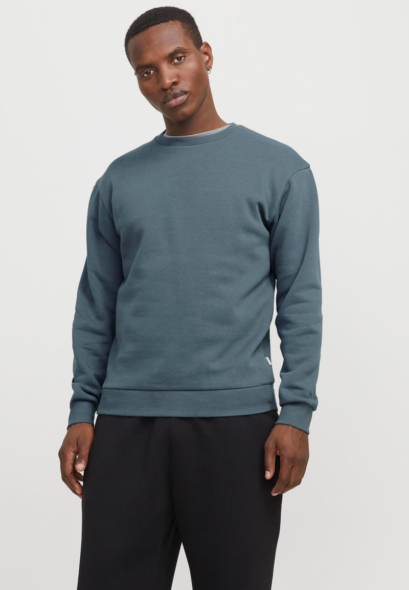 Hombre vestido con un sudadera de cuello redondo azul grisáceo de manga larga y pantalones negros, de pie contra un fondo blanco liso.