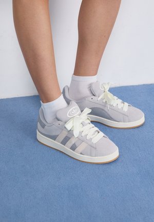 Persona que lleva zapatillas Adidas de color gris claro con cordones y calcetines blancos, de pie sobre una alfombra azul contra una pared blanca.