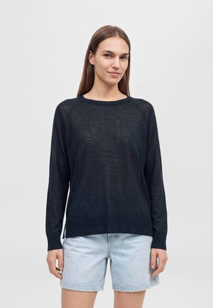 Une femme porte un pull bleu marine foncé à manches longues et un short en jean bleu clair sur un fond uni et clair.