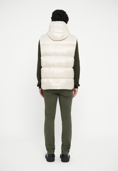 Puffy cream-colored vest with a high collar, worn over a dark green long-sleeve shirt, paired with olive green pants and black shoes.