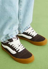 Ténis de lona castanhos com riscas brancas, apresentando uma sola de borracha e atacadores brancos, combinados com jeans de ganga azul claro, sobre uma superfície verde.