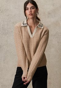 Beige kabelgebreide trui met een diepe V-hals en een witte, gestructureerde kraag met zwarte strepen. Lange, geribde mouwen.