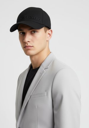 Nuori mies, joka pitää mustaa lippistä ja mustaa paitaa vaaleanharmaan bleiserin alla, katsoo kameraan ilmeettömällä ilmeellä yksivärisellä taustalla.