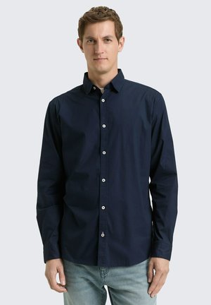 Giovane uomo che indossa una camicia blu marina a maniche lunghe con bottoni e jeans azzurri, in piedi contro uno sfondo chiaro e uniforme.