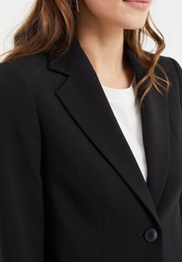 Black blazer with a structured lapel, single button closure, and a smooth texture. Paired with a white top beneath.