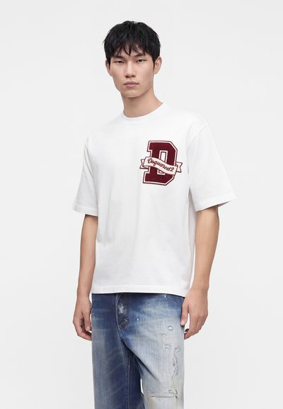 Νέος άνδρας που φοράει λευκό T-shirt με ένα μεγάλο κόκκινο γράμμα D και τζιν με ξεθωριασμένο, φθαρμένο σχέδιο στέκει μπροστά σε ένα απλό φόντο.