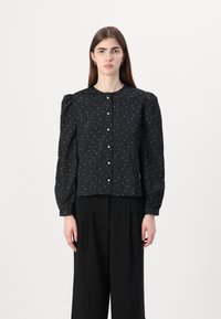 Svart bluse med pufferm, sølvprikker og knappelukking, laget av teksturert stoff. Kombinert med svarte bukser med bred ben.