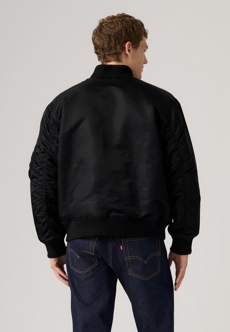 Uomo con giacca bomber nera e jeans blu scuro, visto da dietro su uno sfondo chiaro e uniforme.