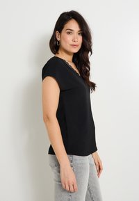 Top negro de manga corta con escote en V, adornado con encaje. Tela texturizada, corte relajado, combinado con jeans gris claro.