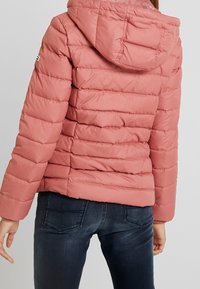 Padded jacket in soft pink, featuring a hood, horizontal seams, and elastic cuffs. Paired with dark jeans, showcasing a casual look.