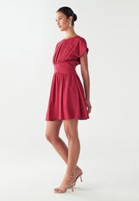 Czerwona krótka sukienka z dopasowanym wcięciem w talii, marszczonym topem i krótkimi rękawami. Posiada rozkloszowaną spódnicę i miękką teksturę. Założona z cielistymi sandałami na obcasie.