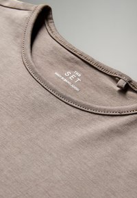 Hnedé bavlnené tričko s okrúhlym výstrihom, hladkou textúrou a prešitými okrajmi. Nálepka hlási "THE SET" a "MADE IN BANGLADESH."