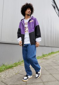 Jachetă ușoară violetă și neagră, tricou alb cu grafice galbene, blugi largați albaștri și adidași negri și albi cu accente galbene.