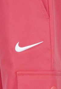 Nike-jakke i lys rosa stoff, med en hvit brodert svosj-logo, lommedetaljer og glatt tekstur.