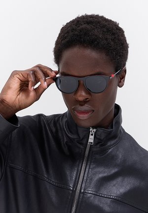 Persona con giacca di pelle nera regola occhiali da sole neri con una mano su uno sfondo semplice.