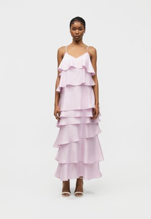 Γυναίκα που φοράει αμάνικο, μακρύ φόρεμα σε ανοιχτό μωβ με στρώσεις από βολάν και λεπτές τιράντες, σε λευκές ανοιχτές γόβες, στέκεται μπροστά σε απλό φόντο.