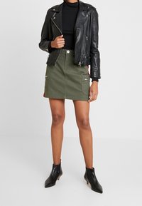 Veste en cuir noir pour motard sur un col roulé noir, associée à une mini-jupe verte olive avec des poches et des détails zippés. Bottines noires.