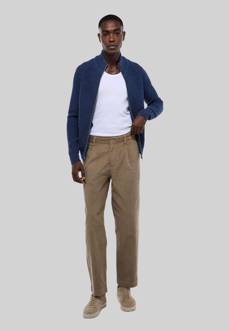 Cardigan zippé en tricot bleu marine porté sur un t-shirt blanc, associé à un pantalon en velours côtelé marron clair et des baskets beiges. Design simple et texturé.