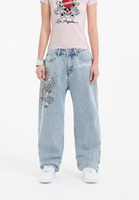 Light blue denim jeans with a relaxed fit, featuring intricate embroidery of a skull and wings, and white branding on the front pocket.
