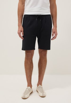 Uomo che indossa pantaloncini neri con coulisse, scarpe da ginnastica bianche e una t-shirt bianca, in piedi davanti a uno sfondo chiaro e uniforme.