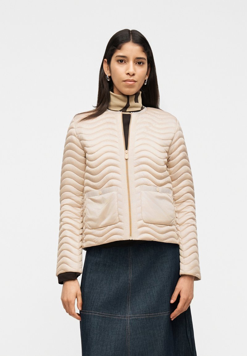 Γυναίκα που φοράει μπεζ καπιτονέ ζακέτα με φερμουάρ πάνω από πουκάμισο με μοτίβο και σκουρόχρωμη τζιν φούστα, στέκεται μπροστά σε απλό λευκό φόντο.