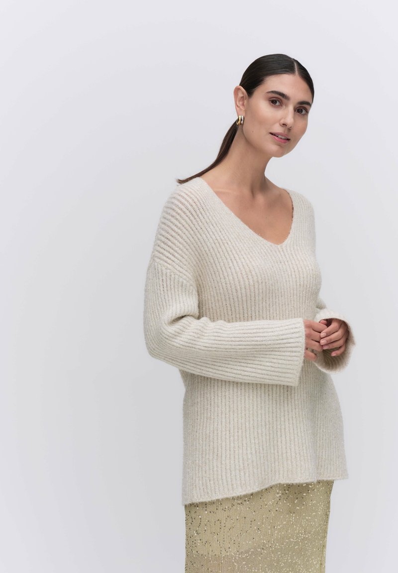 Sweter w kolorze kremowym, z prążkowanego materiału, z dekoltem w kształcie V, luźnymi rękawami i swobodnym krojem, zestawiony z lśniącą złotą spódnicą.