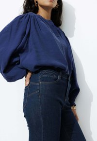 Blouse oversize bleu marine avec des manches bouffantes, en tissu texturé, détails de boutons, associée à un jean foncé ajusté. Accessoires en or simples visibles.