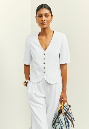 Žena má na sebe biely krátky rukáv s gombíkmi a nohavice, drží modro-bielo pruhovanú kabelku s bambusovými rúčkami.