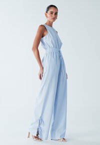 Svetlo modra kombinezon s oprijetim zgornjim delom, elastičnim pasom in širokimi hlačnicami. Ima nastavljive trakove in zlate poudarke na manšetah.