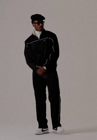 Trening negru cu margini albe, jachetă cu fermoar frontal, pantaloni asortați și adidași albi. Purtați cu o beretă și ochelari de soare.