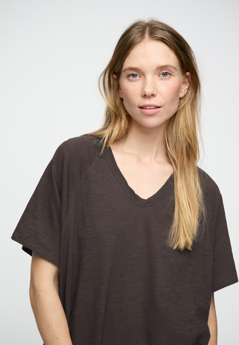 Tamsiai ruda V formos iškirptės marškinėliai su trumpomis rankovėmis ir laisvu siluetu, pagaminti iš lengvos medžiagos su šiek tiek tekstūruota išvaizda.