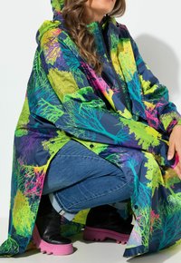 Vrouw in hurkpositie met een kleurrijke regenjas met boompatroon, blauwe spijkerbroek en zwarte laarzen met dikke roze zolen, tegen een witte achtergrond.