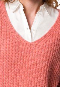 Różowy sweter z dzianiny o dekolcie w serek, z teksturowanym, prążkowanym wzorem, noszony na białej koszuli z kołnierzykiem.