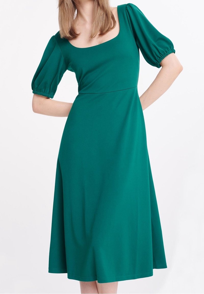 Femme portant une robe bleu sarcelle au genou avec des manches courtes bouffantes et un décolleté carré, debout, mains sur les hanches, sur un fond blanc.