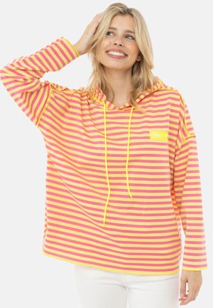 Lächelnde Frau mit blondem Haar, die einen gelb-rosa gestreiften Hoodie und weiße Hose trägt, berührt ihr Haar mit einer Hand.