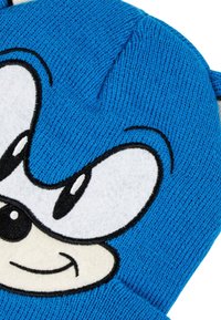 Blauwe gebreide winterhoed met een groot cartoongezicht aan de voorkant, met witte ogen, zwarte pupillen en een lachende mond.