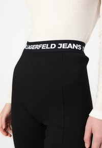 Fekete leggings vastag fekete derékpánttal, amelyen fehér "LAGERFELD JEANS" felirat található. Sima, rugalmas anyag, amely testhez simuló formát biztosít.