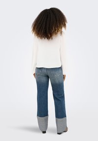 Γυναίκα με σγουρά μαλλιά, ντυμένη με λευκό μπλουζάκι με μακριά μανίκια και τζιν παντελόνι με φαρδιά πόδια, σε διχρωμία, στέκεται με την πλάτη γυρισμένη σε απλό φόντο.