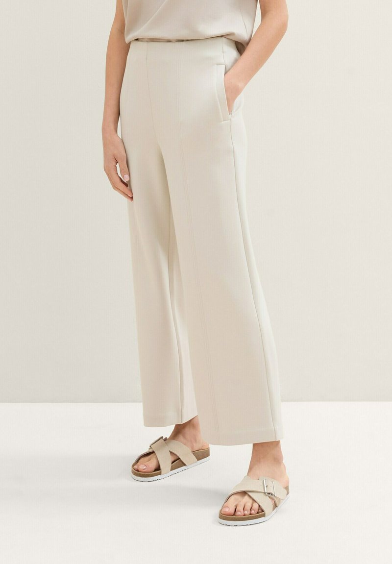 Pantalones de pierna ancha color beige claro con bolsillos laterales; textura suave, líneas limpias y costuras mínimas, combinados con sandalias ligeras con tiras cruzadas.